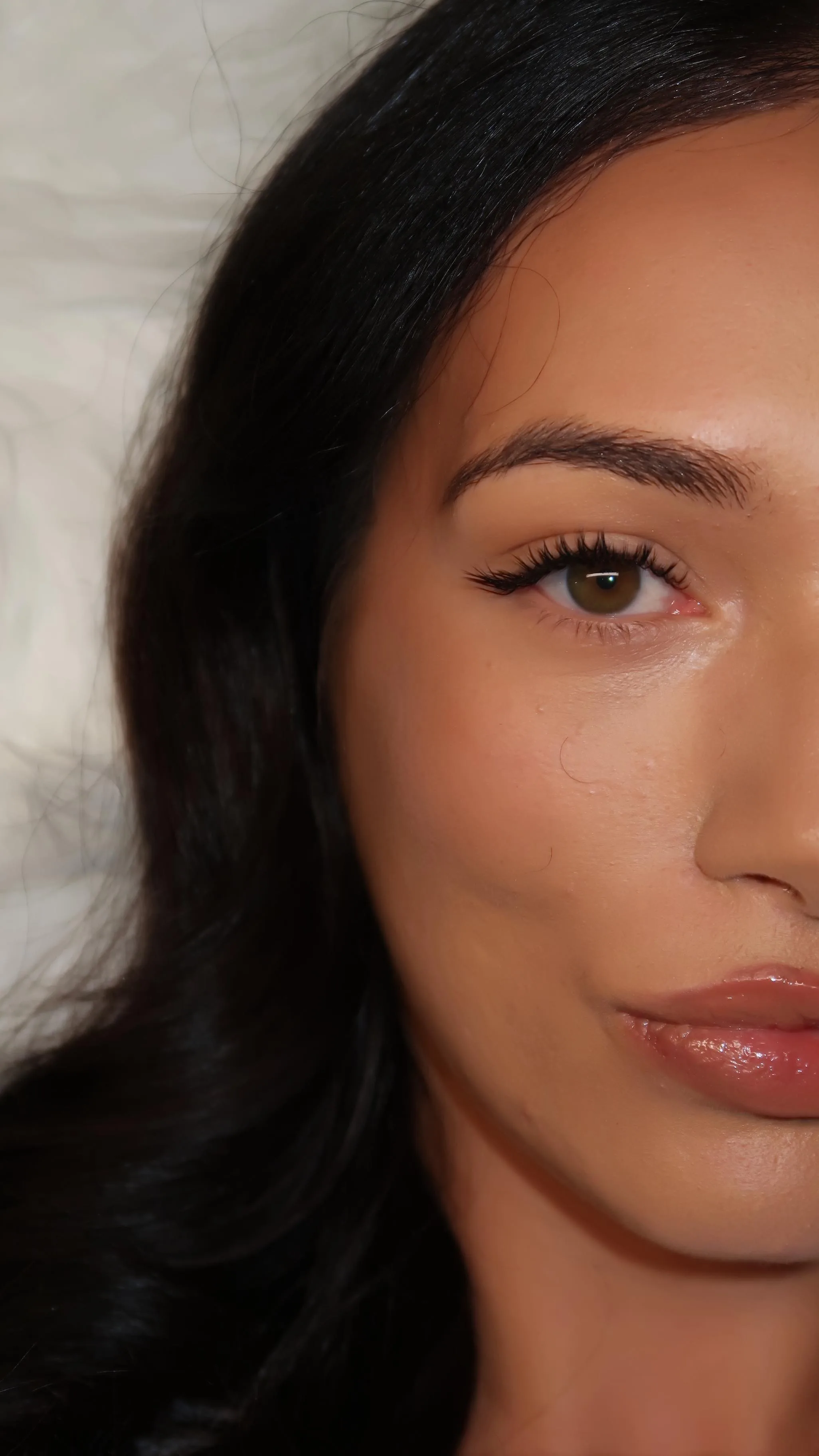 Single brown eye with natural wispy lashes, dark hair, glossy lip
